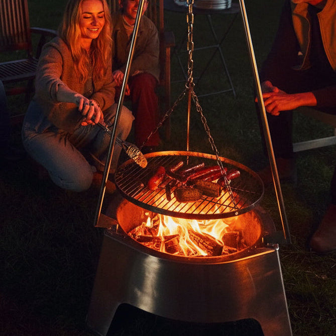 petromax-fire-center-fc1000-station-feuerschale-mit-dreibein-haengerost-bei-lebensieschoen-kaufen_ mit brennenden feuer und grillgut auf dem grillrost